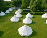 Wioska namiotów typu bell, idealna na glamping. Kilka osób spaceruje po zielonej trawie między białymi, stożkowymi konstrukcjami.