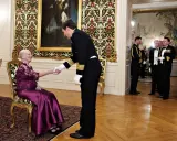 Drottning Margrethe, trots att hon är sjuk, tar emot en officer med ett leende. Hon sitter i en elegant stol, klädd i en lila klänning.