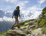 Donna con zaino blu e outfit montagna estate, cammina su sentiero roccioso con cime innevate sullo sfondo.