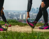 Zwei Läuferinnen dehnen sich auf einem Feldweg mit Blick auf eine Stadt. Sie tragen schwarze Laufhosen und auffällige pink-schwarze Laufschuhe. Ein Zeichen für den Wiedereinstieg ins joggen nach Geburt.