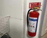 Röd brandsläckare hänger på en vit vägg, redo att användas om brandlarm ljuder. Bredvid en vit metallkorg.