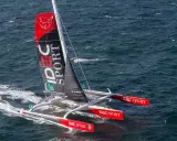 Le trimaran IDEC SPORT fend les vagues, une voile rouge et noire ornée d'un oiseau blanc.