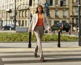 Elegancka kobieta w beżowych stylizacjach zimowych przechodzi przez pasy. Jasny płaszcz narzucony na pomarańczową bluzkę i spodnie w prążki tworzą szykowny miejski look.