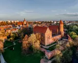Zachód słońca nad zamkami krzyżackimi na Mazurach. Potężne ceglane mury i wieże w blasku złota, otoczone zielenią i miastem.