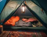 Wnętrze namiotu gotowe do spania. Dwa śpiwory, pomarańczowy i brązowy, czekają na noc. Lampion rzuca ciepłe światło.