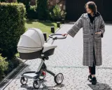 Kobieta w płaszczu prowadzi spacerówkę kubelkową po brukowanej ścieżce wśród zieleni.