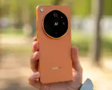 Pomarańczowy oppo flagowiec z charakterystycznym modułem aparatu w dłoni, na tle rozmytego, zielonego krajobrazu.
