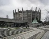 Monumentalny budynek w stylu brutalizm z betonowymi żebrami i szklanym wejściem. Schody prowadzą do wejścia, a wokół rosną drzewa.