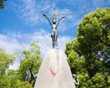 La bambina di Hiroshima, simbolo di pace, con le braccia tese verso il cielo, circondata dal verde degli alberi e dall'azzurro delle nuvole.