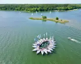 Flota białych żaglówek tworzy okrąg na zielonych jeziorach mazurskich. W tle las i mała wyspa.