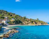 En pittoresk kustby på Elba, Italien, med vita hus som klättrar uppför en grönskande kulle. Klart turkost vatten sköljer mot en sandstrand.
