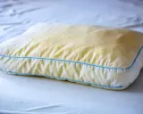 Ein vergilbtes Kissen mit blauen Nähten, das auf einem weißen Bett liegt. Gelbe Flecken deuten auf möglichen Schimmel hin.