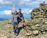 Två glada vandrare njuter av utsikten från en bergstopp under en vandring i Sverige. En stenröse och en modern träkonstruktion med solpaneler syns i bakgrunden.