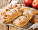 Chrupiące ciasto francuskie z jabłkami, posypane cukrem pudrem, czeka na spróbowanie. Obok leżą rumiane jabłka.