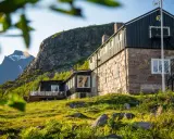 STF Kebnekaise fjällstation med stenfasad och mörk träpanel, belägen vid foten av ett berg. Grönskande sluttning i förgrunden.