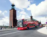 Röd dubbeldäckare med "Hop-on Hop-off Stockholm Buses" på väg förbi Stadshuset. Perfekt för stockholm turistinformation.