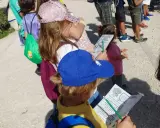 Niños con gorras y mochilas, algunos con folletos, explorando el **parque de Valdebebas** en un día soleado.