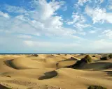 Paisaje de dunas doradas en España, con el mar azul al fondo y un cielo despejado con nubes blancas.