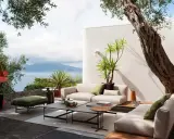 Arredare un terrazzo scoperto con divani chiari, tavolini bassi e un pouf verde, con vista mare e un ulivo secolare.