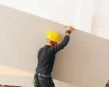 Obrero con casco amarillo transporta una placa de yeso, mostrando varios tipos de yeso apilados.