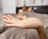 Mano sostiene un condón. En el fondo, una persona en la cama. La decisión de tener sexo oral sin condón es personal.