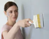 Une femme effectue une retouche peinture murale avec un pinceau gris. Une tache de peinture grise est visible sur sa joue.