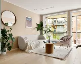 Jasne meble do salonu: puszysta sofa, okrągły stolik, aksamitny fotel i puf. Duże okno z widokiem na balkon.