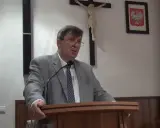 Mężczyzna w garniturze przemawia, za nim krzyż i godło Polski. Może mówi o cnotach kardynalnych.