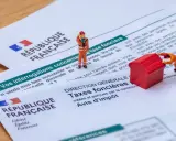 Figurines sur des documents de la République Française concernant la taxe foncière. L'une pousse une maison rouge, l'autre semble réfléchir.