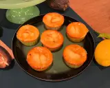 Sechs Zitronenmuffins mit glänzender Glasur auf einem Teller, daneben frische Zitronen und eine Kakaofrucht. Ein leckeres zitronenmuffins rezept!