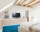 Schlafzimmer im Dachgeschoss, ideal zum Dachboden ausbauen. Helle Farben, Holzbalken und ein blauer Teppich schaffen Gemütlichkeit.