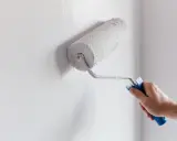 Ręka maluje ścianę wałkiem, nakładając białą farbę. To prosty sposób, jak zamalować farbę ceramiczną, by odświeżyć wnętrze.