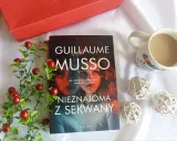 Książka "Nieznajoma z Sekwany" Guillaume'a Musso leży obok kubka kawy i czerwonych ozdobnych kulek.