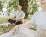 Två personer sitter i lotusställning på gräsmattan under ett träd. De utövar liggande meditation i en lugn parkmiljö.