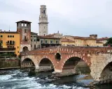 En vacker tegelbro korsar en flod i Verona, Italien. Färgglada hus och ett högt klocktorn syns i bakgrunden.