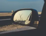 Widok z lusterka fotochromatycznego: droga z latarniami, budynki w oddali i morze.