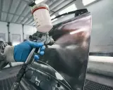 Ręka w niebieskiej rękawiczce trzyma pistolet lakierniczy, rozpylając farbę na element karoserii. Czas schnięcia szpachli na epoksyd jest kluczowy dla dalszych prac.