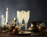 Två eleganta glas med en uppfriskande drink med gin och prosecco, dekorerade med citronskal. Stämningsfull belysning med ljus och ljusslingor skapar en festlig atmosfär.