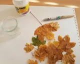 Préparation d'une décoration avec des feuilles d'arbre séchées, de la colle UHU, un pinceau et un petit récipient en verre.
