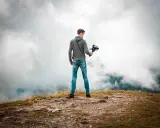 Joven en la cima de una montaña, sosteniendo un **gimbal** para estabilizar su cámara y capturar la vista entre las nubes.