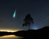 Un **obiettivo per fotografare le stelle** cattura una scia luminosa nel cielo notturno, con un albero solitario in silhouette e un lago calmo.