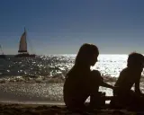 Siluetas de la gomera con niños jugando en la arena, barcos navegando en el mar brillante.