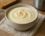 Un bol de béchamel sans beurre, onctueuse et saupoudrée de poivre, prête à être utilisée. Un fouet et une planche à découper sont visibles.
