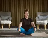 Kvinna i meditationsställning framför en vägg med träribbor. Två fluffiga fåtöljer står på var sin sida. Kanske hon funderar på hur hon ska dekorera hylla i sitt hem.