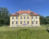 Piękny, żółty pałac w Choroszczy z czerwonym dachem, otoczony zielenią i błękitnym niebem.