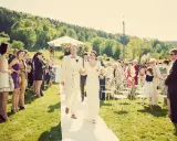 Szczęśliwa para młoda idzie alejką podczas ślubu w lipcu. Goście stoją po bokach, otoczeni zielenią i słońcem.