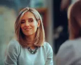 Frau mit kinnlangen Haaren zwinkert und lächelt. Sie trägt einen grauen Pullover und eine auffällige Halskette.