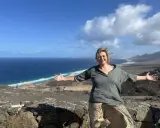 Donna sorridente con le braccia aperte su un paesaggio costiero, un'espressione di gioia che contrasta la solitudine interiore.