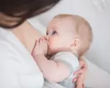 Niemowlę karmione piersią, z niebieskimi oczami, ssie mleko. Czas karmienia jest różny, ale to chwila bliskości.