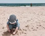 Kleines Kind mit Hut spielt im Sand am Strand der Algarve. Im Hintergrund läuft ein Mann ins Meer. Ein perfekter Tag für Algarve mit Kindern.
